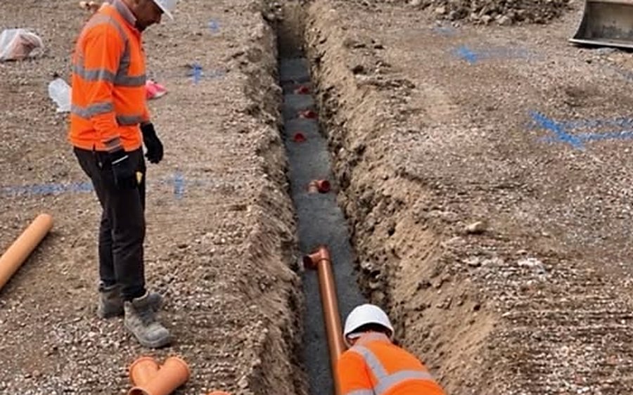 Two operatives in hi-vis laying orange drainage pipe on a gravel bed in a trench